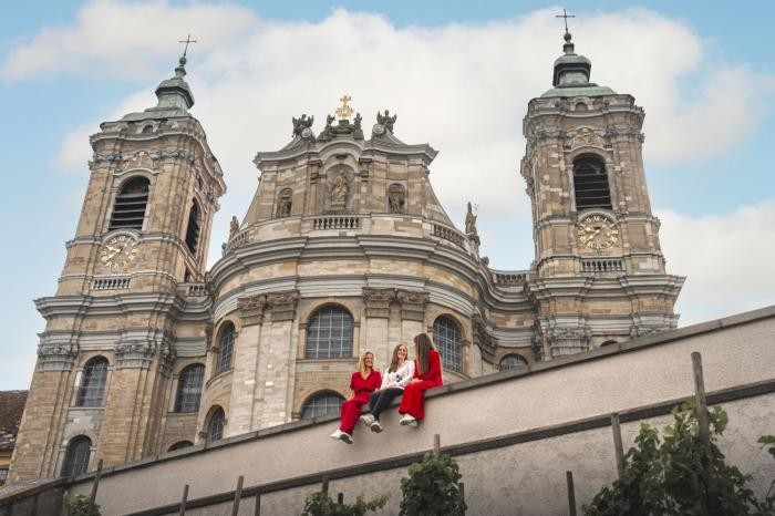 Die drei Mitarbeiterinnen des Stadtmarketings sitzen rot-weiß gekleidet auf der Mauer der Basilika. Im Hintergrund erhebt sich das große Gotteshaus.